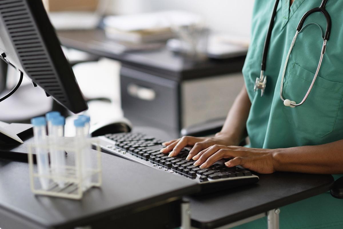  Health care professional wearing scrubs and a stethoscope typing on a computer desktop.