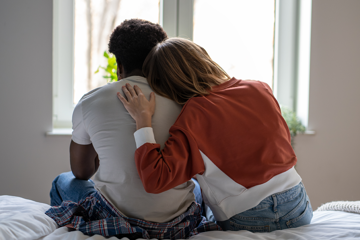 Two people sit on a bed, one leaning on another in a comforting embrace